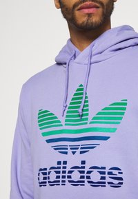 Lila hoodie med en framträdande grön och blå randig Adidas trefoil-logotyp samt matchande text, tillverkad av ett mjukt tyg.