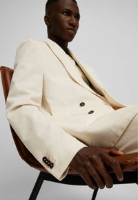 Cremefarbene Kordhosen-Doppelreiher-Anzug mit braunen Knöpfen, getragen im Sitzen auf einem braunen Ledersessel; glatte Textur und strukturiertes Design.