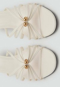 Sandales ouvertes en crème avec plusieurs fines lanières formant un motif de nœud, présentant deux perles décoratives centrales en or et une semelle noire.