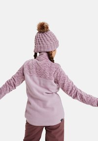 Helle rosa Fleecejacke mit gemusterten Ärmeln, hohem Kragen und abgerundetem Saum. Begleitet von einer gestrickten rosa Mütze mit einem Pompon.