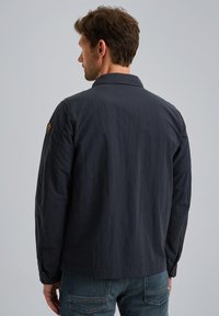 Marineblaue, leichte Jacke mit langen Ärmeln, geknöpften Manschetten und einem Kragen. Verfügt über ein Abzeichen auf der linken Schulter und eine glatte Textur.