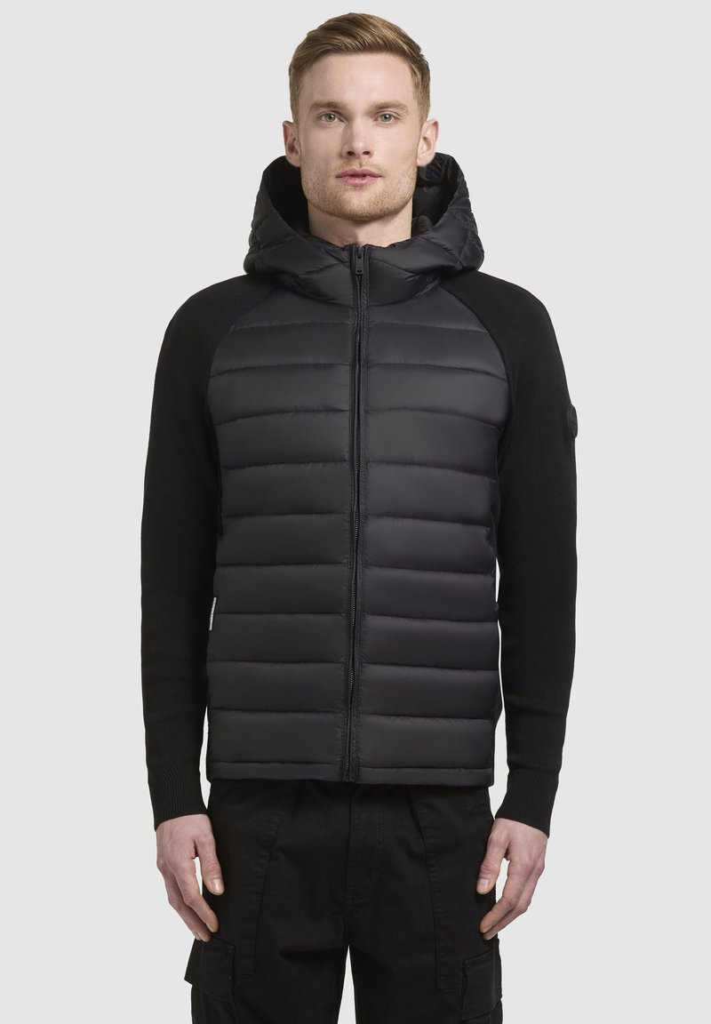 Giacca a vento nera con design a maglia a coste, chiusura con zip e cappuccio. Presenta pannelli a trapunta orizzontali. Dettagli in tessuto misto.