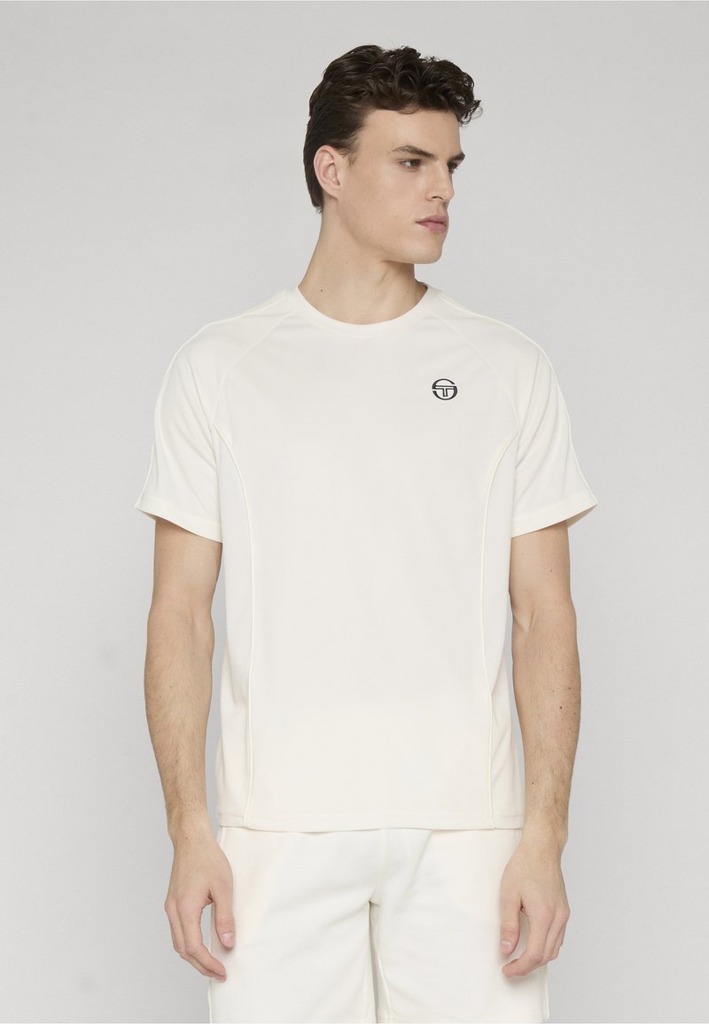 Jeune homme portant un t-shirt blanc uni à manches courtes avec un petit logo noir sur le côté gauche de la poitrine, tourné vers la droite sur un fond clair.