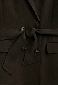 Blazer marron foncé avec ceinture à nouer, deux poches avant et boutons noirs. Tissu élégant avec une texture lisse et un design ajusté.