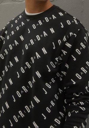 Person iført en sort sweatshirt med hvidt diagonalt "JORDAN" tekstmønster, der dækker stoffet, synlig fra nakke til midt på overkroppen.