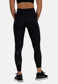 Czarne legginsy z wysokim stanem, gładką teksturą i kanciastymi szarymi wzorami po bokach. W zestawie z czarnymi butami sportowymi.