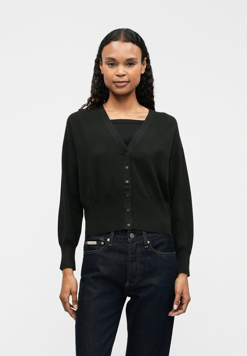 Zwarte, korte cardigan met een v-hals, lange mouwen en knoopsluiting. Draagt over een zwarte top, in combinatie met donkere jeans. Soepele textuur.