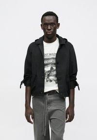 Čierna ľahká bunda s kapucňou a nastaviteľnými rukávmi, oblečená cez biele tričko s potlačou, v kombinácii so sivými nohavicami s opaskom.