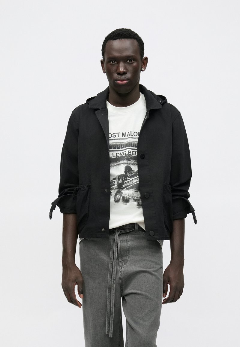 Čierna ľahká bunda s kapucňou a nastaviteľnými rukávmi, oblečená cez biele tričko s potlačou, v kombinácii so sivými nohavicami s opaskom.