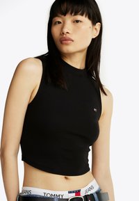 Sort cropped tanktop med høj hals, fremstillet af ribbet stof. Har et lille logo på brystet og bæres med et bælte, der viser branding.