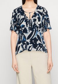 Blusa de estampado floral en azul marino y celeste, con mangas cortas con volante, escote de ojo de cerradura y tejido ligero. Combinada con pantalones beiges.