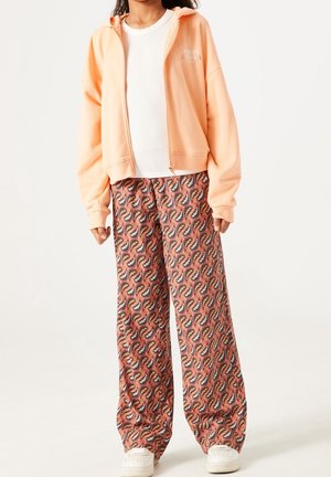 Person wearing peach zip-up hoodie, white ribbed shirt, wide-leg pants with orange and black paisley pattern, and white sneakers.