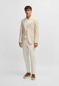 Käynyt vaaleanbeige pellavablazeri, jossa on kaksi etutaskua, yhdistettynä valkoiseen pyöreäkaulaiseen paitaan ja kermaisiin housuihin, viimeistelty ruskeilla mokkasneakereilla.