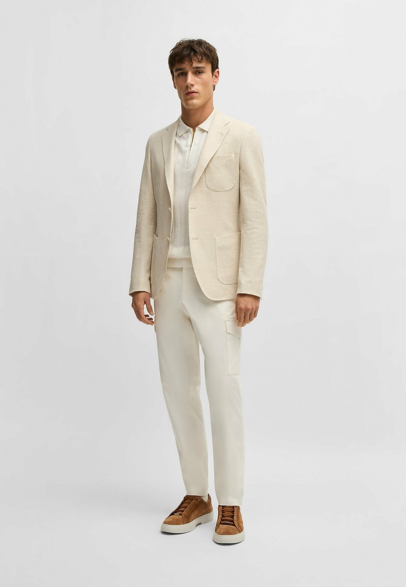 Käynyt vaaleanbeige pellavablazeri, jossa on kaksi etutaskua, yhdistettynä valkoiseen pyöreäkaulaiseen paitaan ja kermaisiin housuihin, viimeistelty ruskeilla mokkasneakereilla.