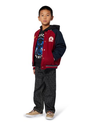 Rode en marineblauwe varsityjack met patches, zwarte grafische hoodie, gepatternde zwarte broek en beige sneakers met dierenontwerpen.