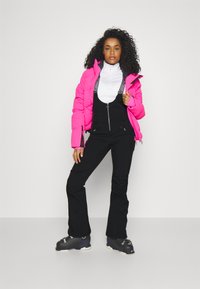 Helle rosa isolierte Jacke, die über einem weißen Langarmoberteil getragen wird, kombiniert mit schwarzen Skihosen und schwarzen Skistiefeln. Einfaches Design und kontrastierende Farben.