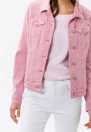 Kvinde iført en lyserød denimjakke, lyserød ribstrikket top og hvide bukser mod en ensfarvet hvid baggrund.
