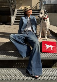 Denimjakke og vide jeans i lys blå. Svarte platåsandaler. Dalmatinerhund som sitter i nærheten. Rød kunstverk med en hvit hund.