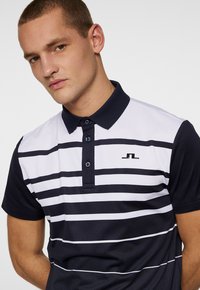 Marinblå och vit randig polo med krage, med tre knappar och en logotyp på bröstet. Mjuk textur och figurnära design.