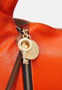 Borsa in pelle arancione con un charm a forma di chiave dorata e un'etichetta rotonda con la scritta "by Chloé" in rilievo. Zip con un elemento in tessuto nero.