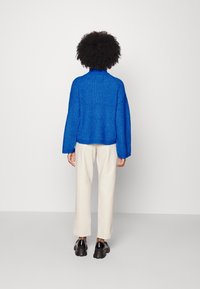 Blå stickad tröja med en avslappnad passform och hög halsringning, kombinerad med krämfärgade byxor och svarta kraftiga skor. Synlig texturerad mönster.