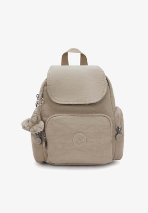 Beżowy plecak z przodem i bocznymi kieszeniami na zamek, górną klapą, tkaninową rączką oraz małym pluszowym breloczkiem w kształcie małpki przymocowanym do bocznego zamka.