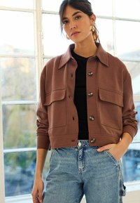 Braune, cropped Jacke mit einem Knopfverschluss, zwei großen Brusttaschen und einem klassischen Kragen, kombiniert mit hellblauen, hoch taillierten Jeans.
