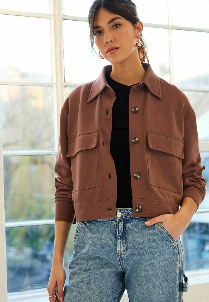Braune, cropped Jacke mit einem Knopfverschluss, zwei großen Brusttaschen und einem klassischen Kragen, kombiniert mit hellblauen, hoch taillierten Jeans.