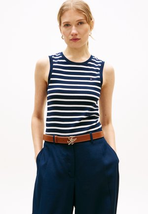 Femme portant un haut sans manches rayé bleu marine et blanc, un pantalon bleu marine et une ceinture marron avec des initiales dorées, debout les mains dans les poches.