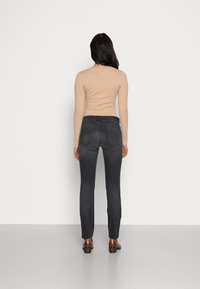 En åtsittande, ribbad beige långärmad tröja i kombination med mörkgrå jeans, sedd bakifrån, med bruna skor och en slät golvyta som bakgrund.