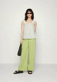 Blusa branca sem mangas com detalhe franzido, combinada com calças largas verde claro. Os acessórios incluem uma bolsa tecida preta e sandálias.