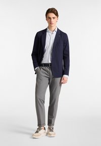 Marineblazer, lichtblauw overhemd, grijze broek en beige sneakers. De blazer heeft een zak; het shirt heeft een gestreept patroon; de broek is op maat gemaakt.