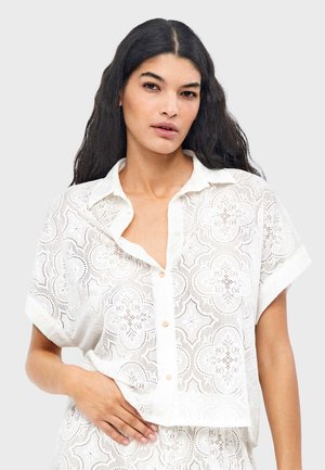 Joven con cabello largo y oscuro que lleva una camisa blanca de manga corta con botones y encaje con patrones florales.