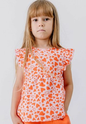 Young girl with long blonde hair wearing a white dress with orange heart patterns and ruffled sleeves, standing against a plain white background.