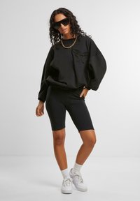 Svart, oversized hettegenser med preget grafikk, kombinert med svarte sykkelshorts og hvite joggesko, med ribbestrikkede mansjetter og krage.