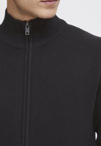 Svart stickad tröja med hög krage och full dragkedja. Har ribbad struktur och en slim fit-design som framhäver nackområdet.