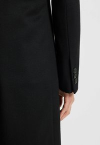 Cappotto di lana nero con una texture liscia, caratterizzato da un fit su misura, maniche lunghe e tre bottoni rotondi sul polsino.