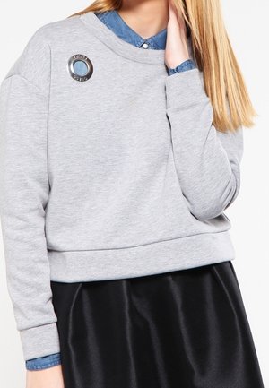 Jonge persoon draagt een grijze trui met een ronde metalen speld op de linkerschouder, gedragen over een blauw overhemd met kraag en een zwarte rok.