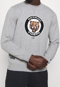 Šedá mikina s ribovými manžetami a pasem, na přední straně s grafickým logo Detroit Tigers. Viditelná texturovaná tkanina.
