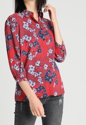 Woman wearing a red floral blouse with three-quarter sleeves and dark grey jeans against a plain background.