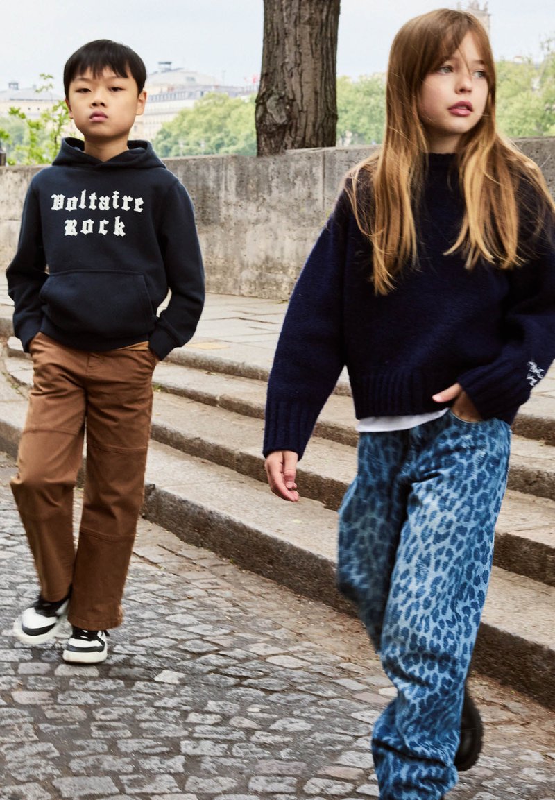 Twee kinderen lopen op een stenen pad met stappen; de een draagt een zwarte "Voltaire Rock" hoodie en een paarse broek, de ander een marineblauwe trui en een blauwe broek met luipaardprint.