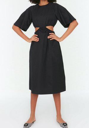 Femme portant une robe midi noire avec des détails découpés à la taille, des manches larges et courtes, et des talons à carreaux avec une boucle décorative.