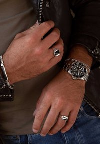 Silver watch with black face, chain bracelet, and angular silver ring on a hand. Textured leather jacket, grey shirt, and blue jeans visible.