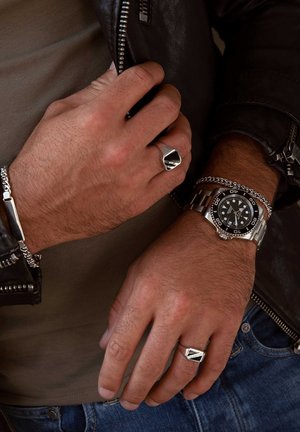 Silver watch with black face, chain bracelet, and angular silver ring on a hand. Textured leather jacket, grey shirt, and blue jeans visible.