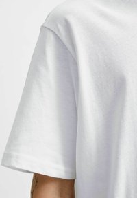 Balta kokvilnas T-kreklu ar apaļu kaklu un īsām piedurknēm, ar mīkstu tekstūru un tīru, minimālistisku dizainu. Redzami šuvju detaļas.