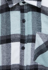 Gros plan sur une chemise en polaire avec un grand motif à carreaux noir, blanc et bleu clair, des boutons noirs, un col et une poche poitrine.