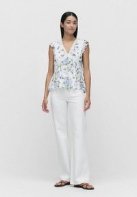 Weißes Blusenshirt mit floralen Mustern, Rüschensäumen, Spitzenverzierung und ausgestelltem Saum, kombiniert mit weißen, weiten Hosen und schwarzen Sandalen.
