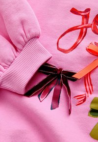 Sweatshirt rose avec des poignets côtelés, orné d'imprimés floraux colorés et de deux accents en ruban—un en orange et un dans une couleur veloutée sombre.