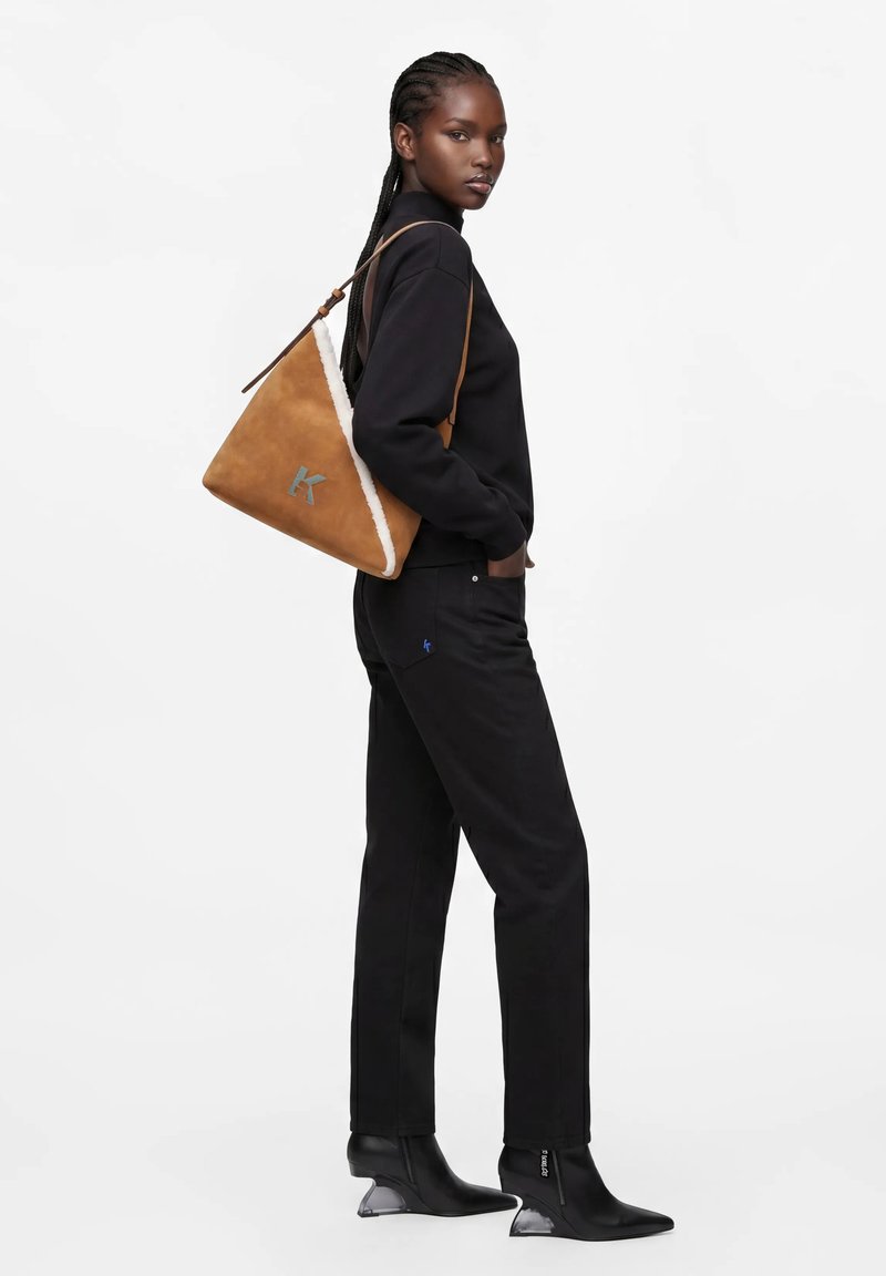 Femme aux cheveux tressés porte un pull noir, un pantalon noir et des bottes à talons noires, portant un sac à épaule triangulaire beige avec une bordure blanche.