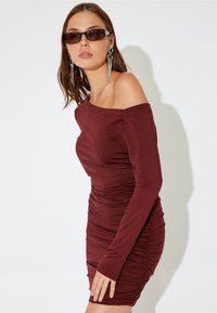 Bordeauxfarbenes Off-Shoulder-Kleid mit langen Ärmeln, gerafftem Stoff, tailliertem Schnitt und glattem Material. Kombiniert mit getönten Sonnenbrillen und auffälligen Ohrringen.